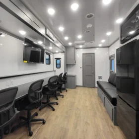 A modern command center trailer interior with a row of office chairs, wall-mounted TV, workstations with outlets, gray cabinetry, and wood-look flooring under bright LED ceiling lights.