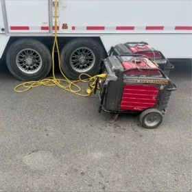 Two portable generators with red casings are connected to a trailer by yellow power cables, parked on asphalt beside dual trailer wheels.