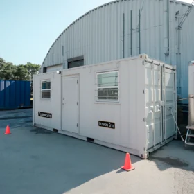 A white ground-level office unit with a "Fusion Site" sign is positioned beside a blue shipping container on a paved area outside a large metal building under a clear blue sky.