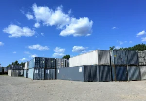 A large open lot with stacks of 40-foot shipping containers in shades of gray and blue under a bright blue sky with scattered white clouds.