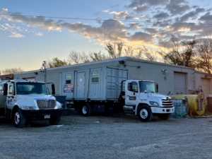 A white service truck and a portable restroom unit are parked on gravel beside a gray building, with a yellow tank and trees visible against a colorful sunset sky.