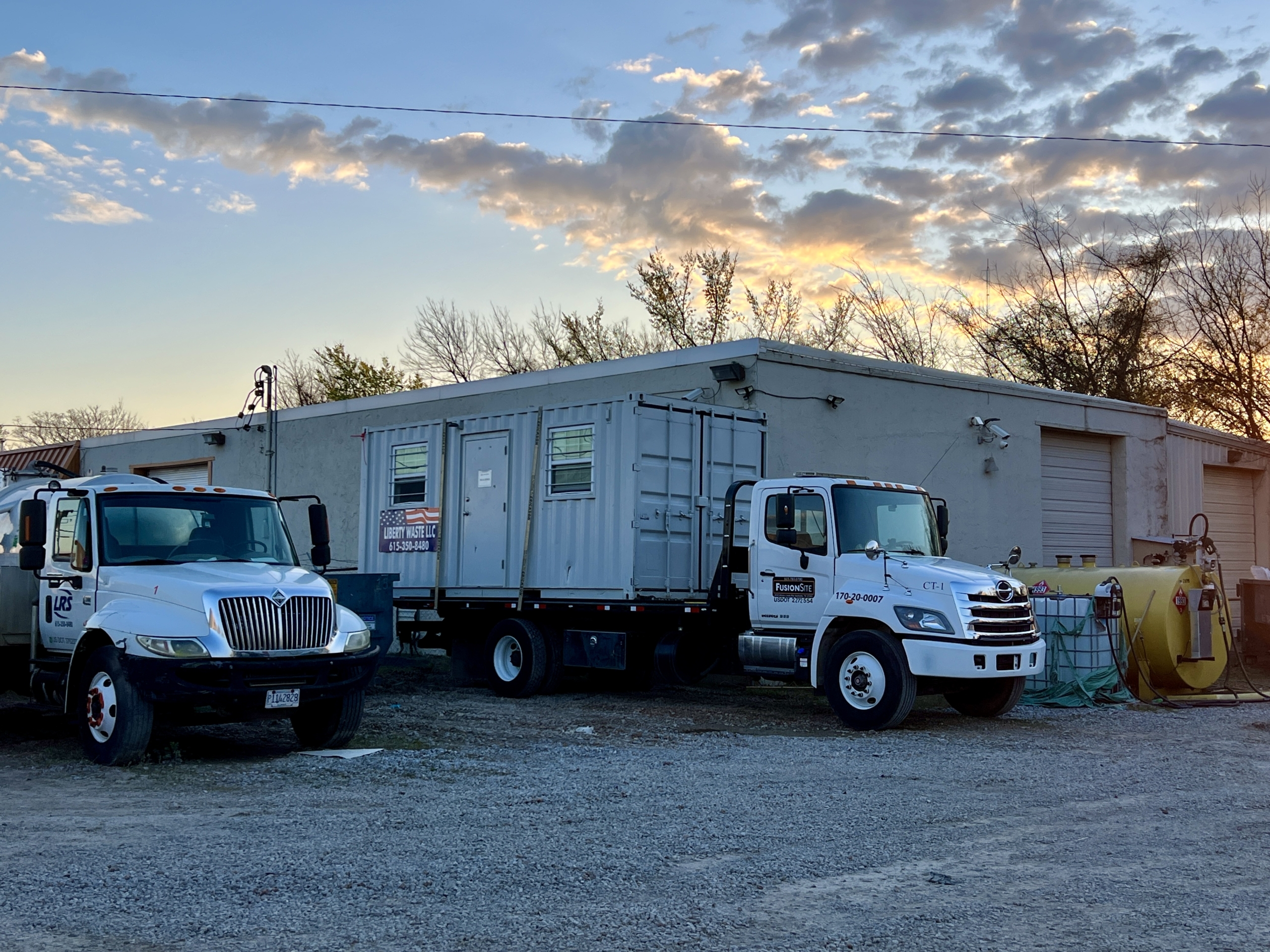 A white service truck and a portable restroom unit are parked on gravel beside a gray building, with a yellow tank and trees visible against a colorful sunset sky.