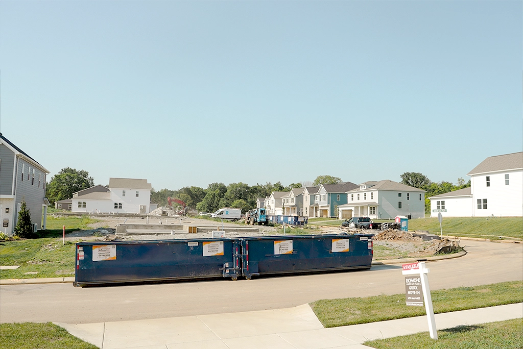 A blue construction dumpster is positioned on a dirt road in front of several partially built houses, with construction equipment visible in the background under a clear blue sky.