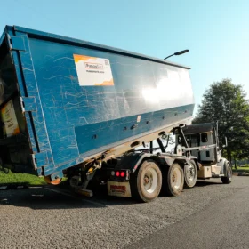 A blue dumpster truck is positioned on a gravel road, tilted to unload waste, with green grass and trees visible in the background under a clear blue sky.