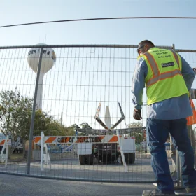 A construction worker in a reflective safety vest stands near a chain-link fence, with a water tower and a work vehicle visible in the background under a clear blue sky.