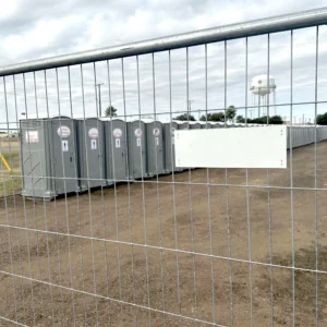 A row of gray portable restrooms is enclosed by a chain-link fence on a gravel lot, with a water tower and power lines visible in the background under a cloudy sky.
