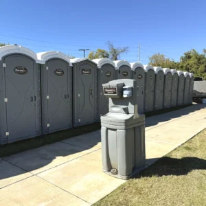 A row of gray portable restrooms is lined up along a concrete path beside a handwashing station, with a trailer parked in the background under a clear blue sky.