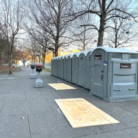 A row of gray portable restrooms is positioned along a paved walkway lined with bare trees and wooden platforms, with a person walking in the distance on a clear day.