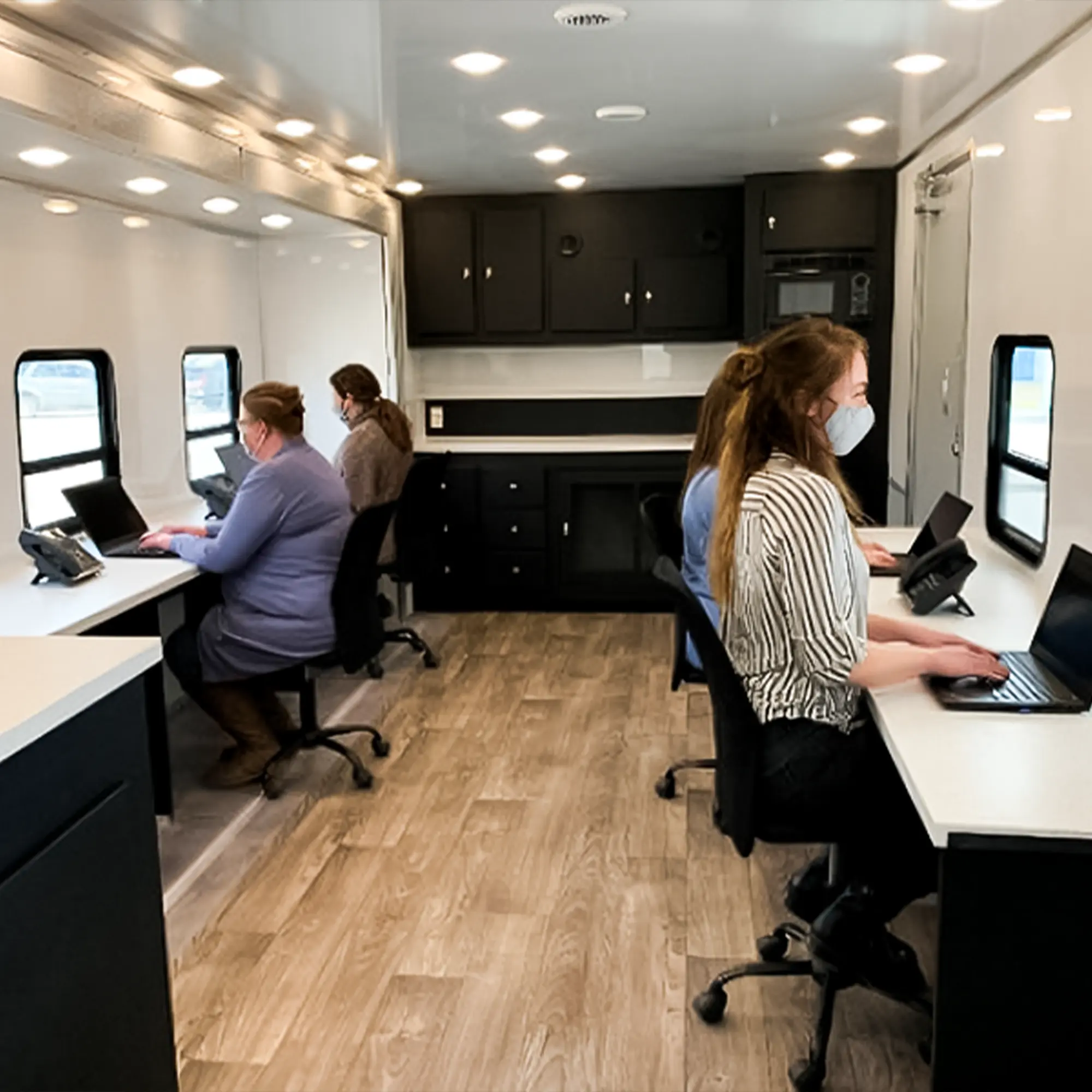 Inside a mobile office trailer, three masked women are seated at desks with laptops, working under bright lights and near windows, with dark cabinetry and a neutral-colored floor.