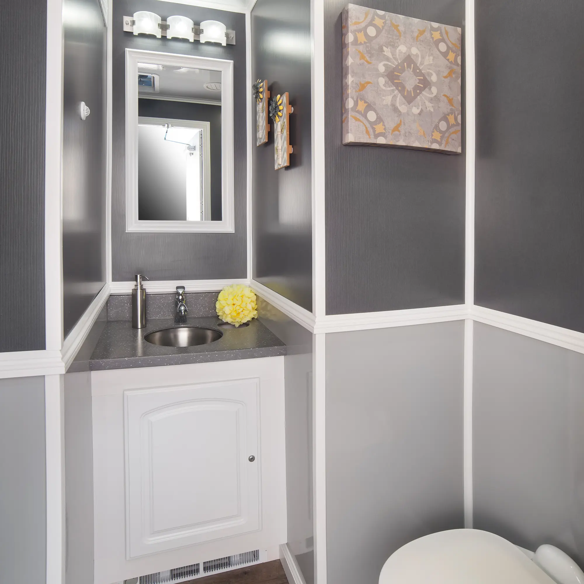 A modern portable restroom interior features a white toilet, a sleek sink with a silver faucet, decorative wall art, and soft lighting, all set against dark gray walls and a wooden floor.