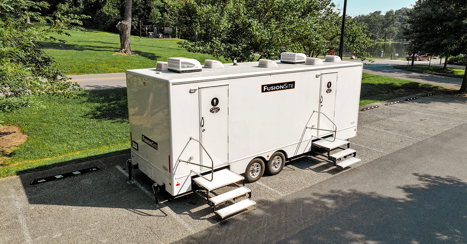 A white portable restroom trailer with multiple entrances is parked on a gravel lot surrounded by trees, with a road and a body of water visible in the background on a sunny day.