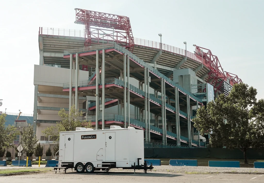 A white portable restroom trailer is parked on a gravel lot beside a large stadium under a clear sky, with concrete structures and trees in the background.