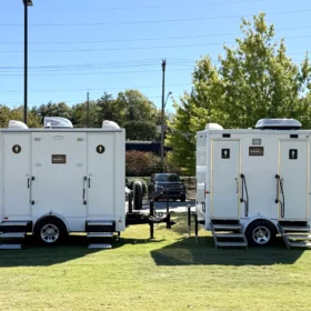 Two white portable restroom trailers are parked on green grass beside a black fence, with a white service truck visible on a paved road in the background under a clear blue sky.