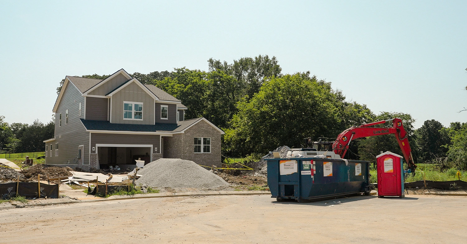 A red portable restroom is positioned beside a blue construction dumpster and a large excavator at a residential building site, with a partially constructed house and piles of gravel in the background.