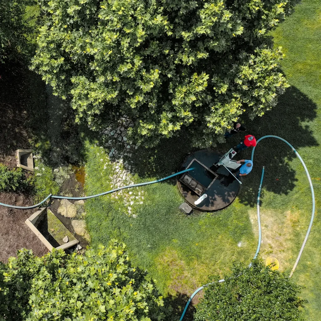 A silver sanitation truck is parked on the side of a road, with a blue hose extending to a nearby access point surrounded by trees and grassy areas on a sunny day.