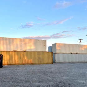 A row of stacked shipping containers in various colors is positioned on a gravel lot under a clear sky, with a blue dumpster nearby and utility poles in the background.