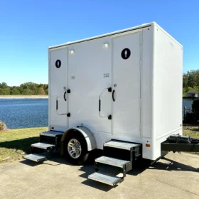 A white restroom trailer with separate men’s and women’s entrances is parked beside a lake on a sunny day, featuring metal steps and handrails for access.