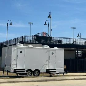 A white restroom trailer is parked outside a university sports stadium with tall light poles and bleachers visible under a clear blue sky.