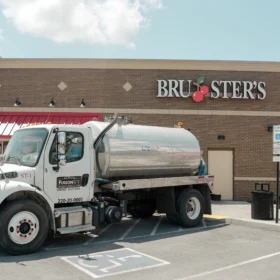 A white sanitation truck with a shiny metal tank is parked in a designated space outside a brick building with a red awning, near a trash can and a reserved parking sign on a sunny day.