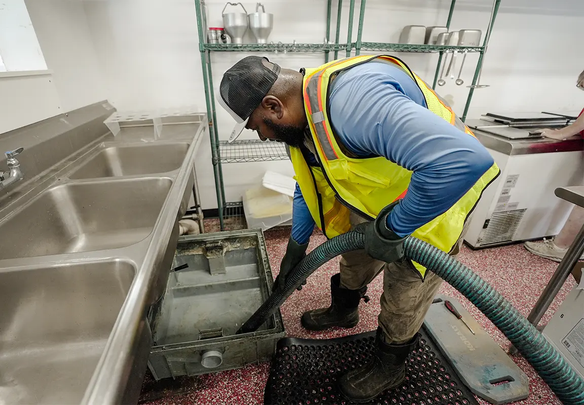 A worker in a yellow safety vest and gloves is using a hose to clean an indoor grease trap in a commercial kitchen, surrounded by stainless steel sinks and storage shelves.