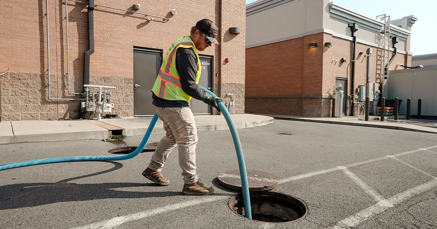 A worker in a reflective vest is using a blue hose to service a manhole in a parking lot beside a brick building, with utility pipes visible on the wall behind him.