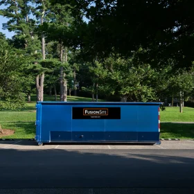 A blue dumpster with company signage is parked on a paved area beside a grassy field and trees under a clear blue sky.