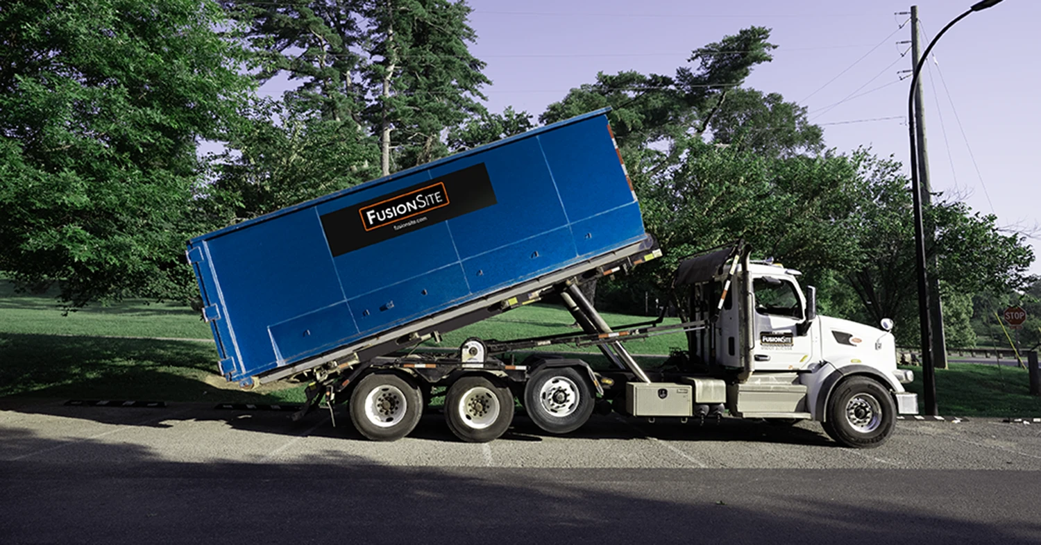 A blue roll-off dumpster is tilted on the back of a white truck, parked on a paved road near a grassy area and a stop sign under a clear blue sky.