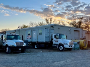 A white service truck and a portable restroom unit are parked on gravel beside a gray building, with a yellow tank and trees visible against a colorful sunset sky.