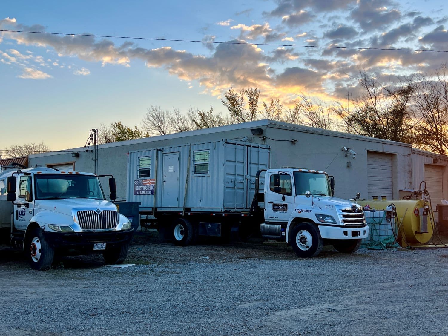 A white service truck and a portable restroom unit are parked on gravel beside a gray building, with a yellow tank and trees visible against a colorful sunset sky.